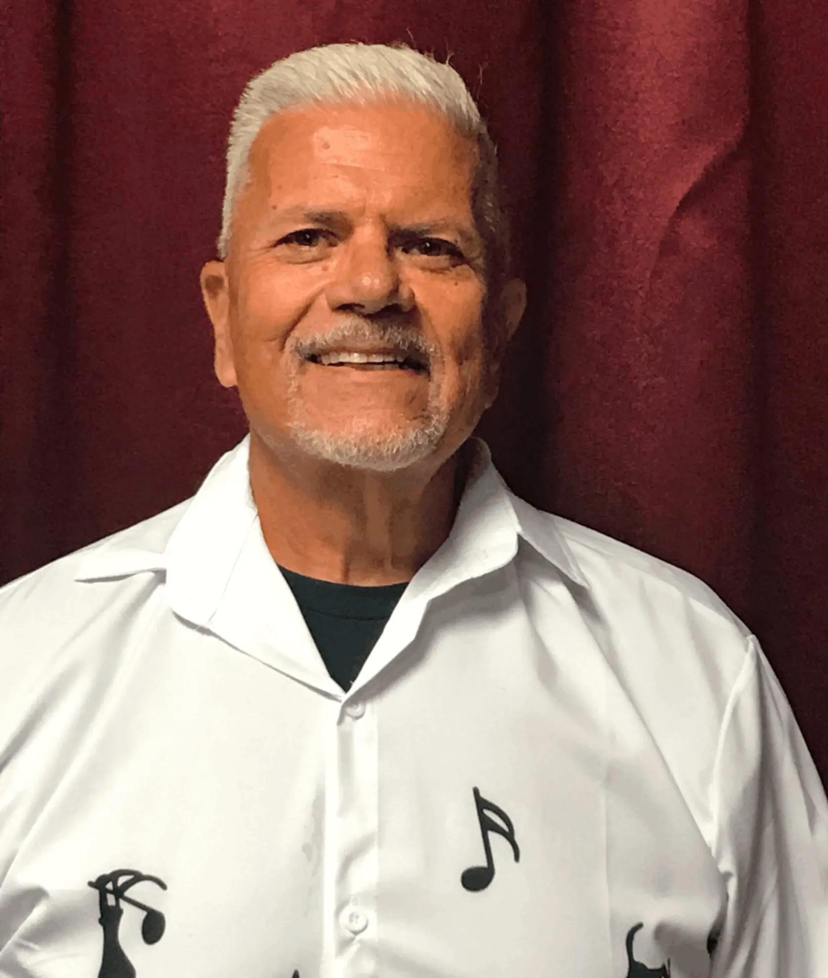 Smiling man wearing a white shirt with musical notes stands against a maroon curtain.