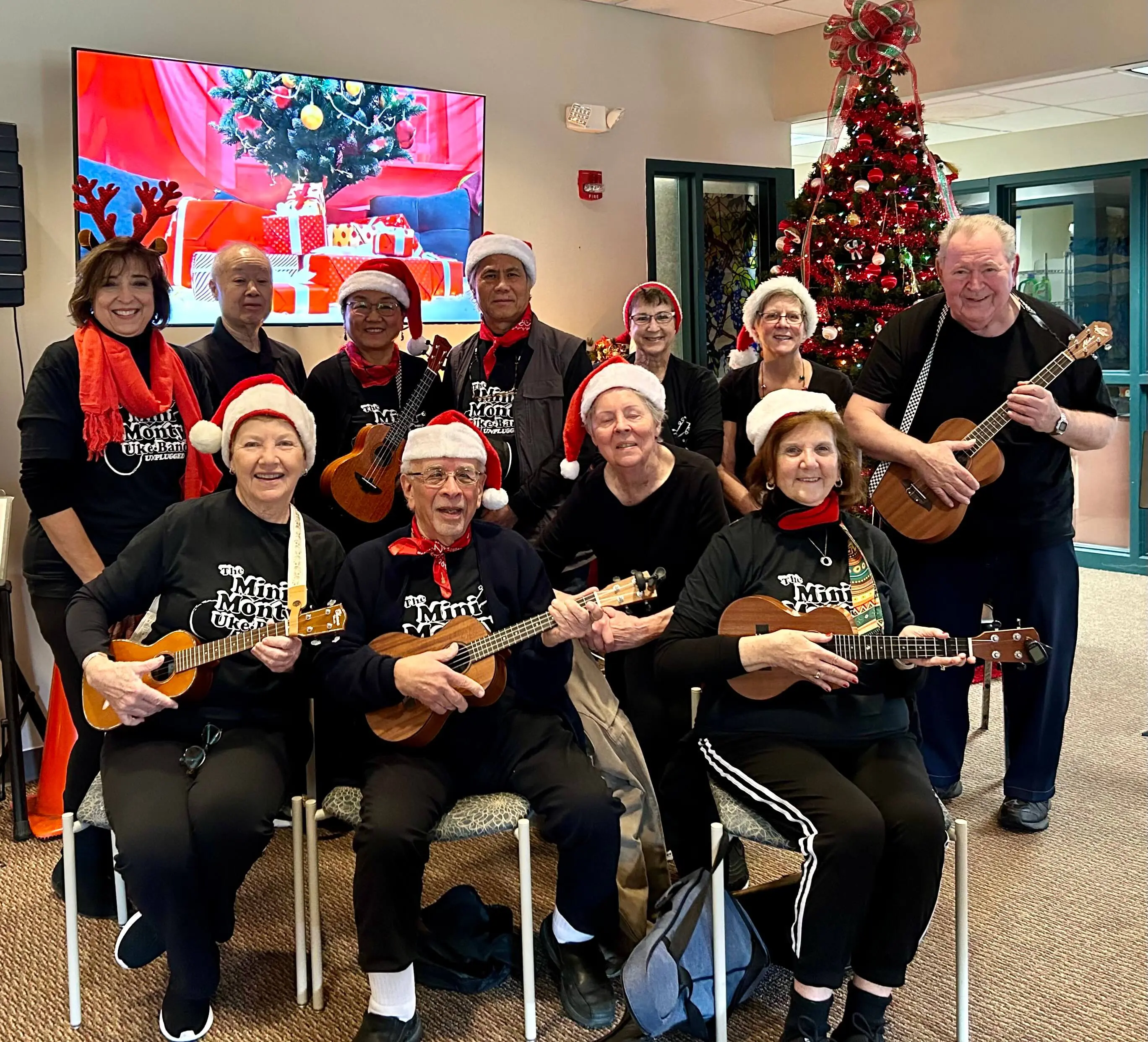 Group wearing Santa hats playing ukuleles in a festive room.