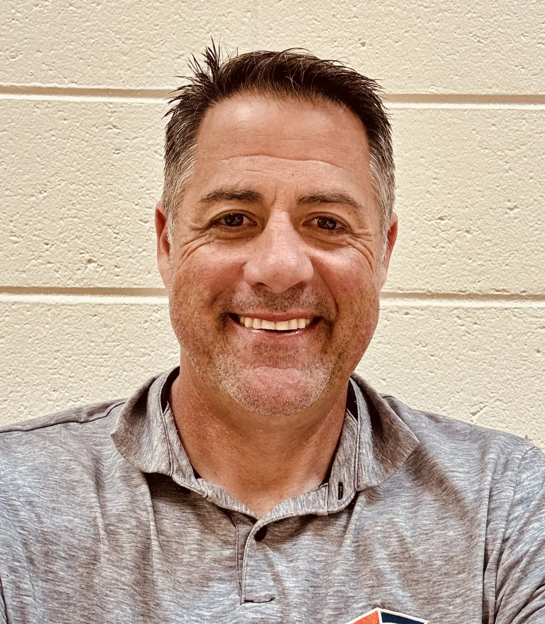 Smiling man in a gray polo shirt standing against a beige wall.